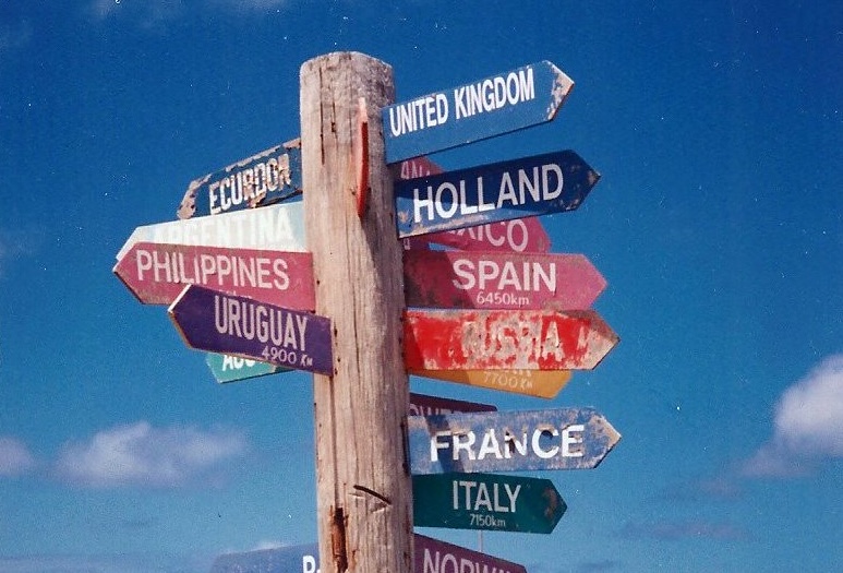 Wooden signpost with colorful directional arrows pointing to different countries including United Kingdom, Holland, Spain, France, Philippines, Uruguay, and others against a blue sky.