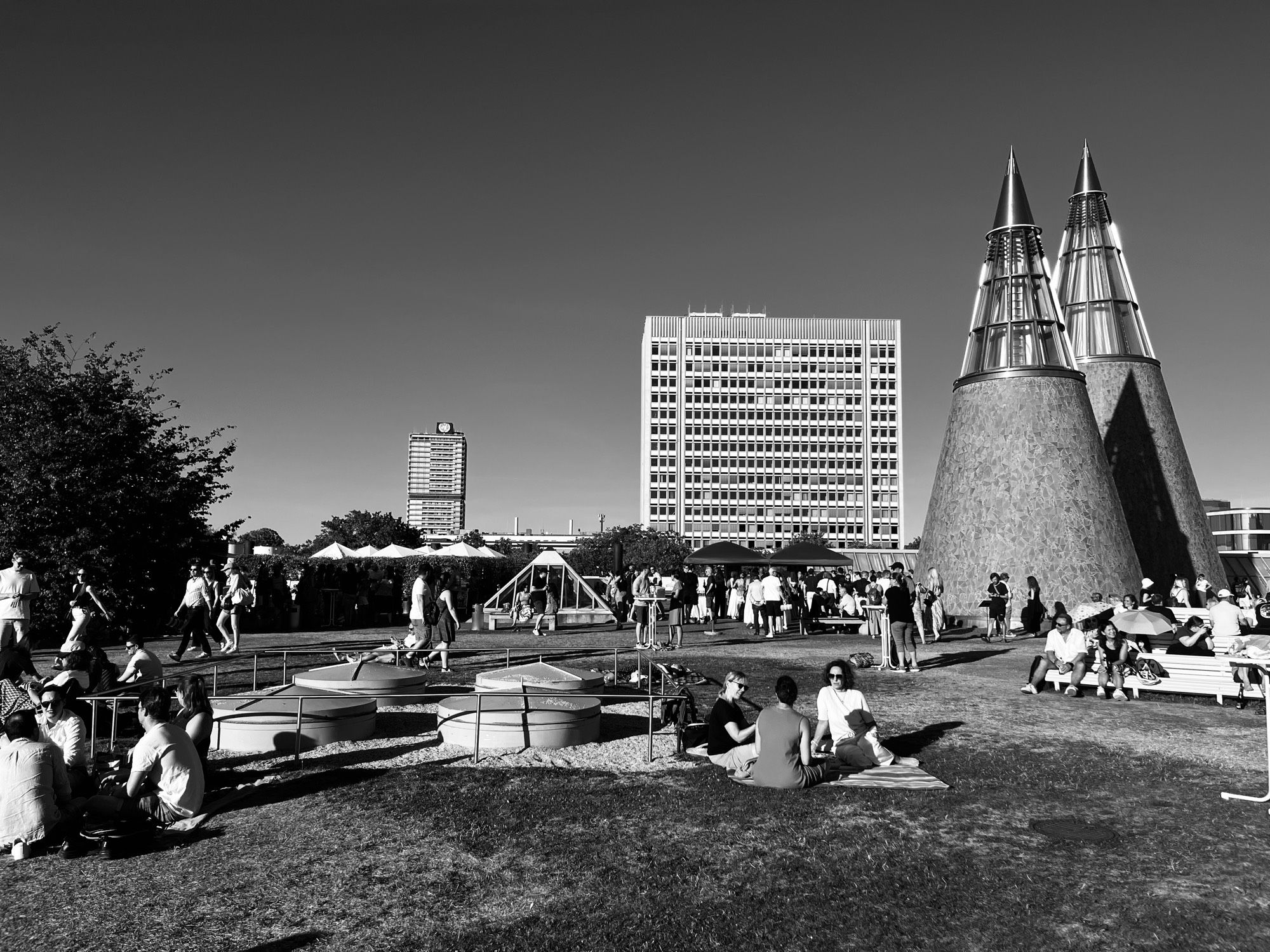 Black and white photograph of a rooftop lawn crowded with people sitting and standing in low evening sun. Two tall glass and stone cones rise at the right. Behind, a wide office block with a grid of windows and a slimmer tower carrying a circular emblem, against a near-black sky.