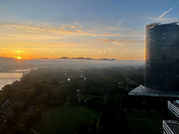 A high-angle morning view at sunrise over Bonn. In the foreground right, the curved glass facade of the Post Tower reflects the morning light. Below, the trees of the Rheinaue and the Rhine river are partially shrouded in a blanket of low-lying morning mist. Across the horizon, the silhouette of the Siebengebirge hills emerges above the fog into a golden and blue sky, with the rising sun just clearing the mist on the left.